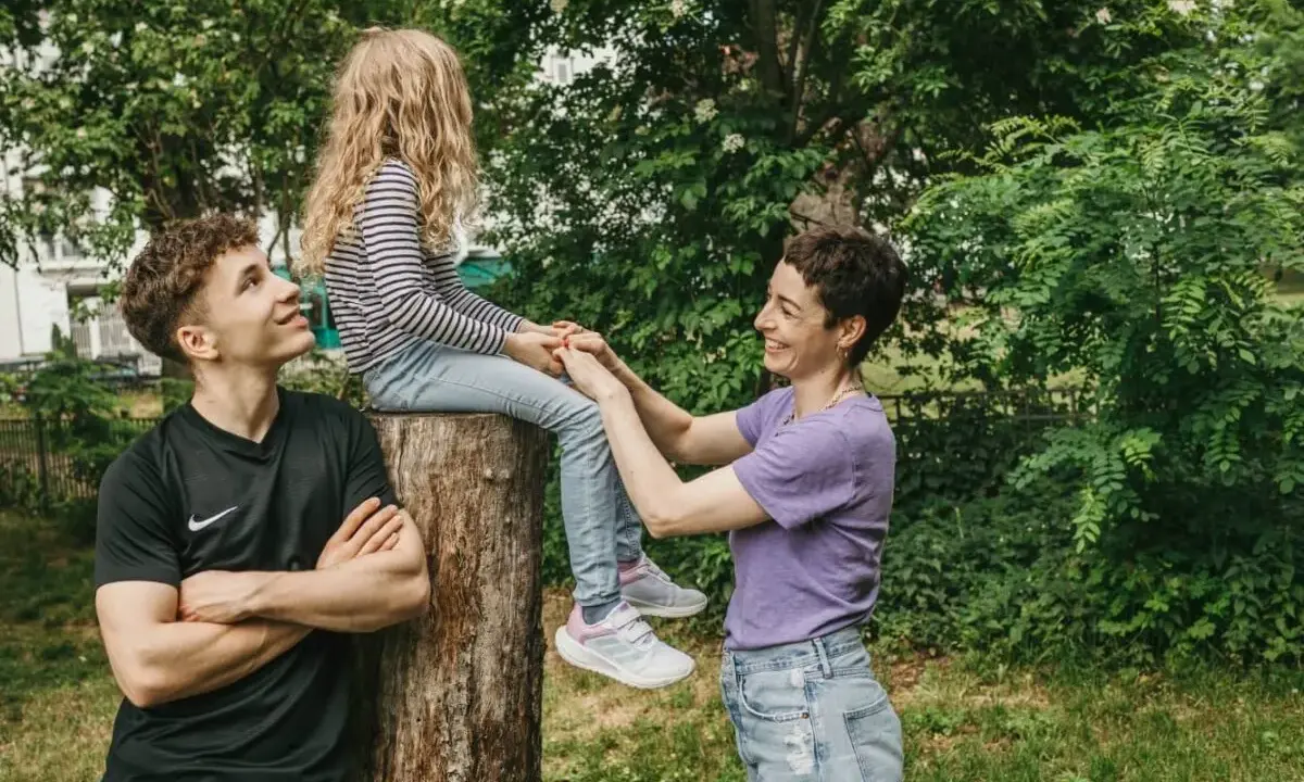 Ein junger Mann schaut zu seiner Mutter, die mit seiner kleinen Schwester auf einem Baumstumpf sitzt. Vielleicht denkt er, wenn der Sohn die falsche Freundin hat, ist die Familie trotzdem da.