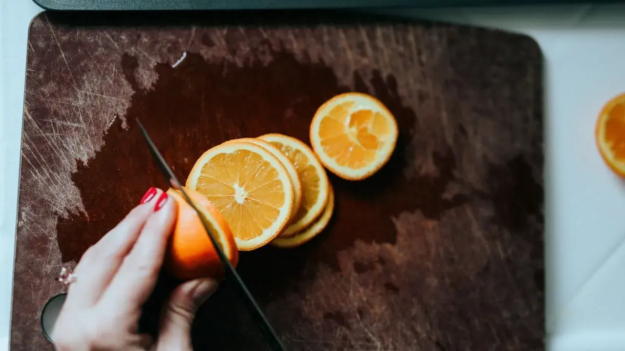 Eine Hand mit roten Nägeln schneidet Orangen auf einem Holzbrett. Orangen in der Schwangerschaft sind eine gesunde Wahl.