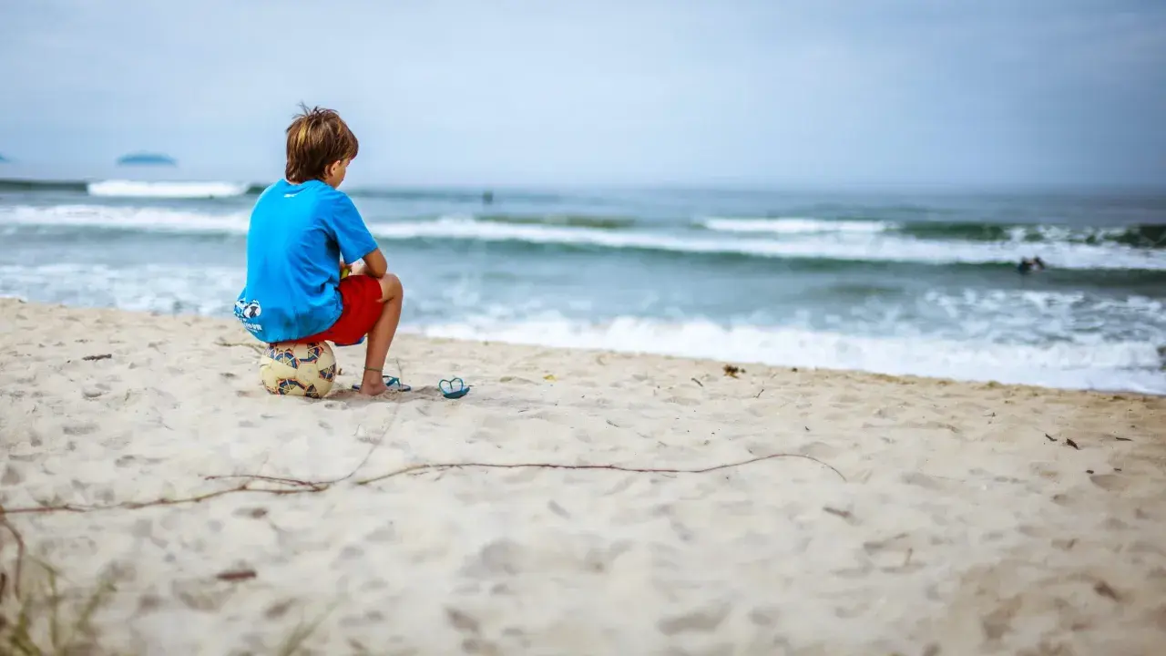 Ein Junge sitzt allein am Strand auf einem Fußball. Er blickt aufs Meer. Meine Tochter 16 hat keine Freunde, sie fühlt sich oft so einsam.