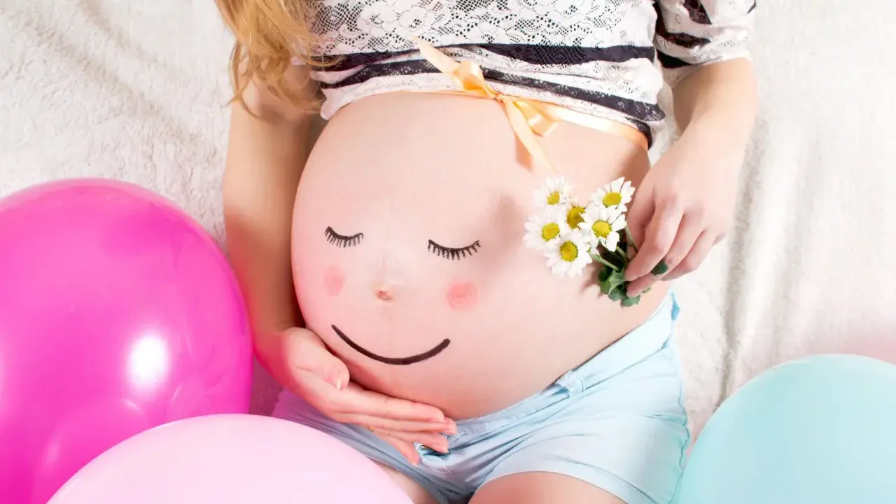 Ein bemalter Babybauch mit einem lächelnden Gesicht, umgeben von bunten Ballons und Gänseblümchen. Ein süßer Moment des Babybauch bemalen.