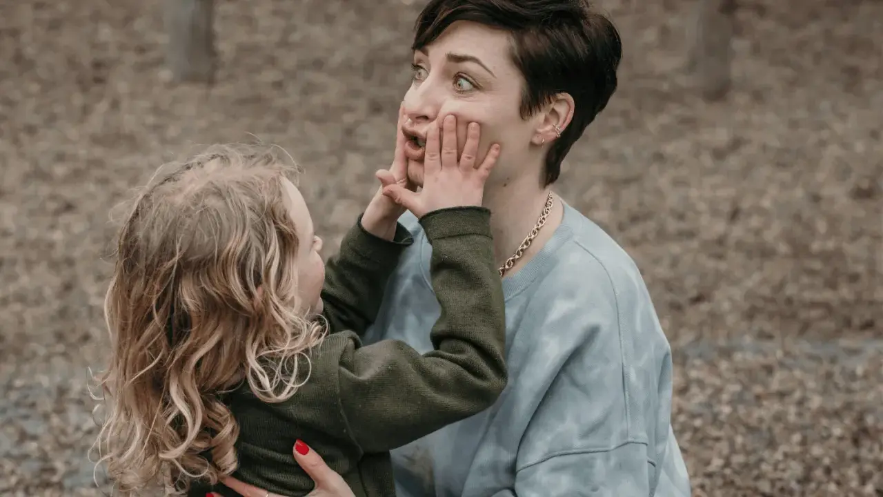 Ein kleines Kind drückt der Mutter spielerisch die Hände ins Gesicht. Vielleicht fragt es sich, warum beleidigen Kinder andere Kinder, wenn sie doch so viel Spaß haben können.