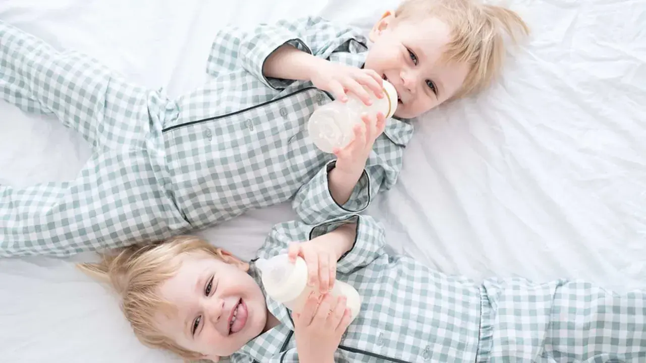 Zwei süße Jungen in karierten Pyjamas trinken aus Fläschchen. Perfekt für Inspiration für Zwillingsnamen Jungen!