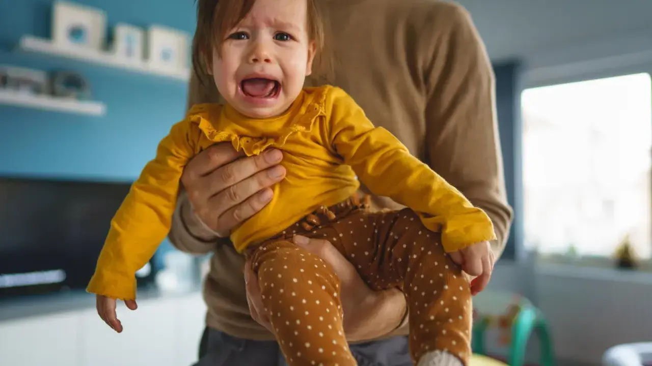 Ein weinendes Baby in gelbem Oberteil und gepunkteter Hose wird von seinem Vater gehalten. Typische "Mama-Phase".