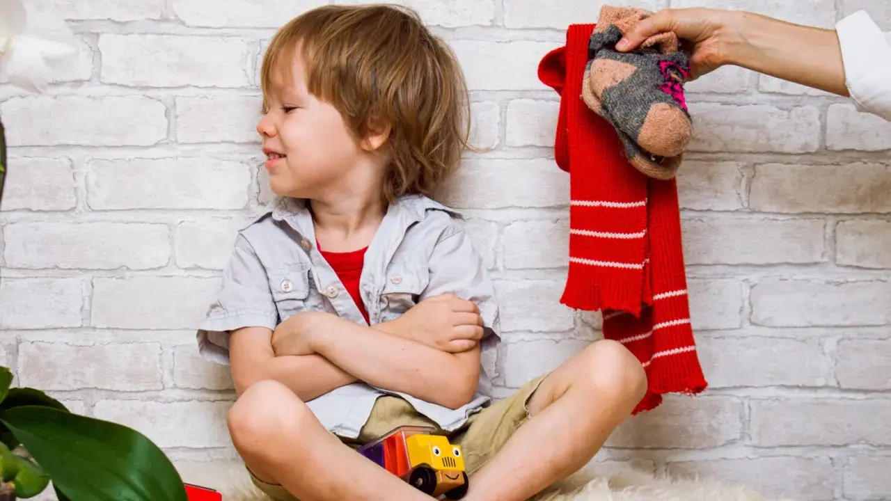 Ein Junge sitzt mit verschränkten Armen und einem trotzigen Gesichtsausdruck da. Über ihm hält eine Hand einen roten Schal und ein Paar Socken. Der Junge will sich nicht anziehen.