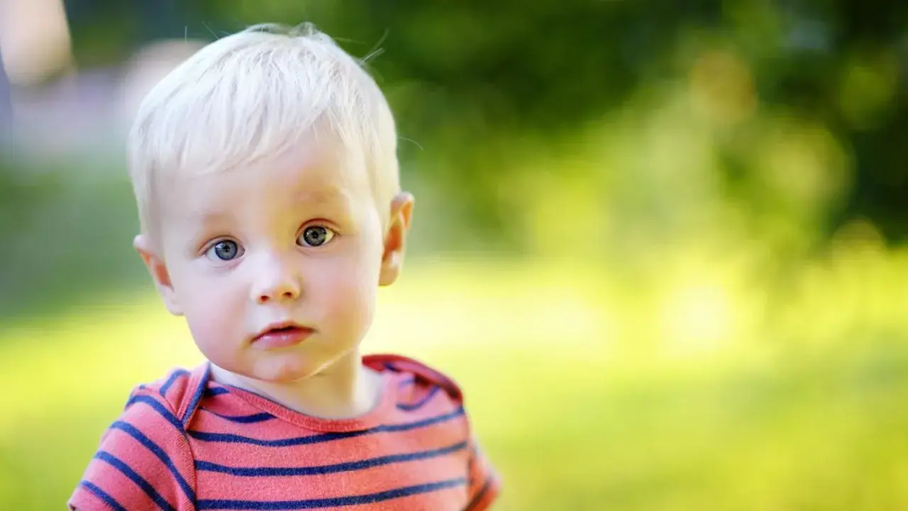 Ein kleiner Junge mit blonden Haaren und blauen Augen blickt neugierig in die Kamera. Sein gestreiftes Shirt erinnert an die Vielfalt von Jungennamen international.