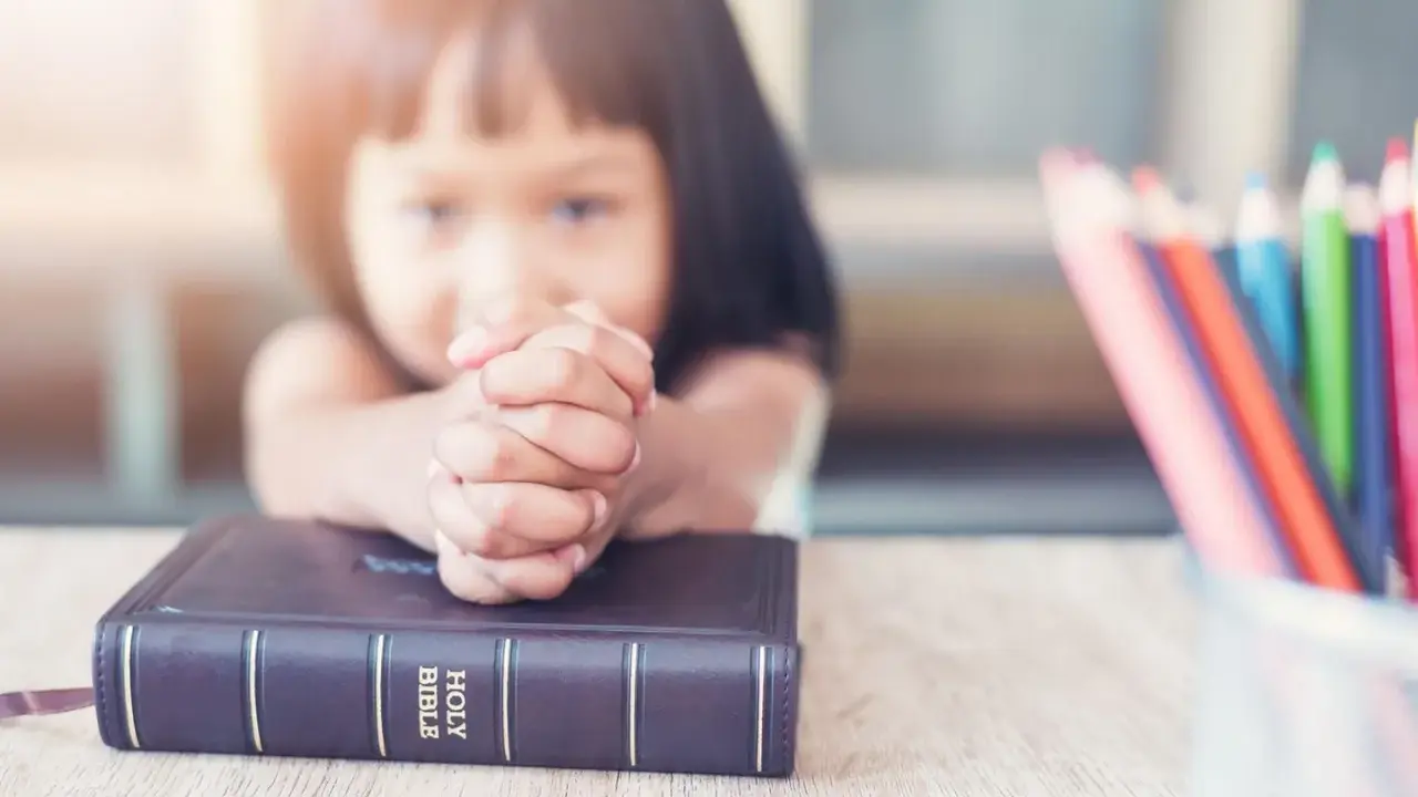 Ein kleines Mädchen betet mit gefalteten Händen über einer Bibel. Vielleicht lernt sie gerade etwas über biblische Frauennamen. Buntstifte liegen daneben.
