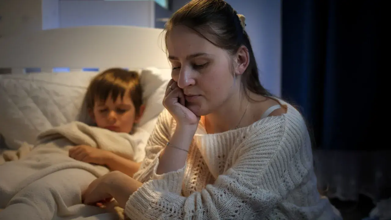 Müde Mutter sitzt neben ihrem schlafenden Sohn, der immer beim Aufwachen weint. Sie hält ihren Kopf in der Hand.