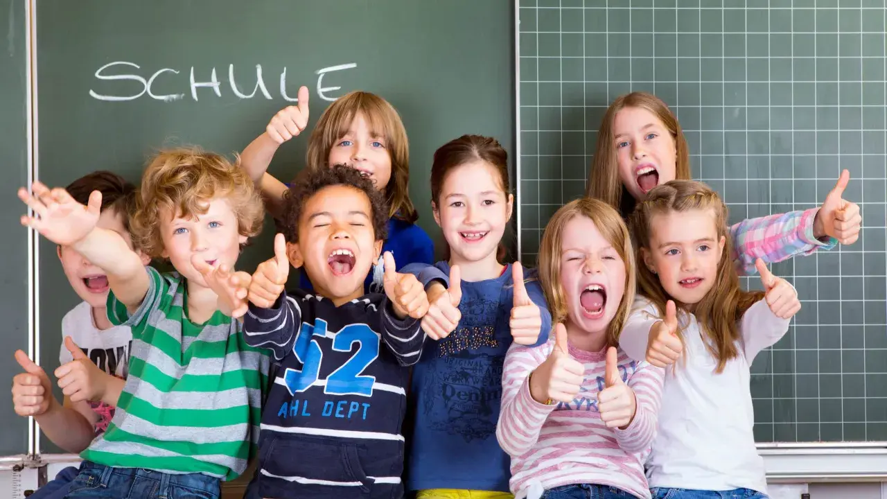 Fröhliche Kinder mit Daumen hoch vor einer Tafel mit dem Wort "SCHULE". Ein perfekter erster schultag!