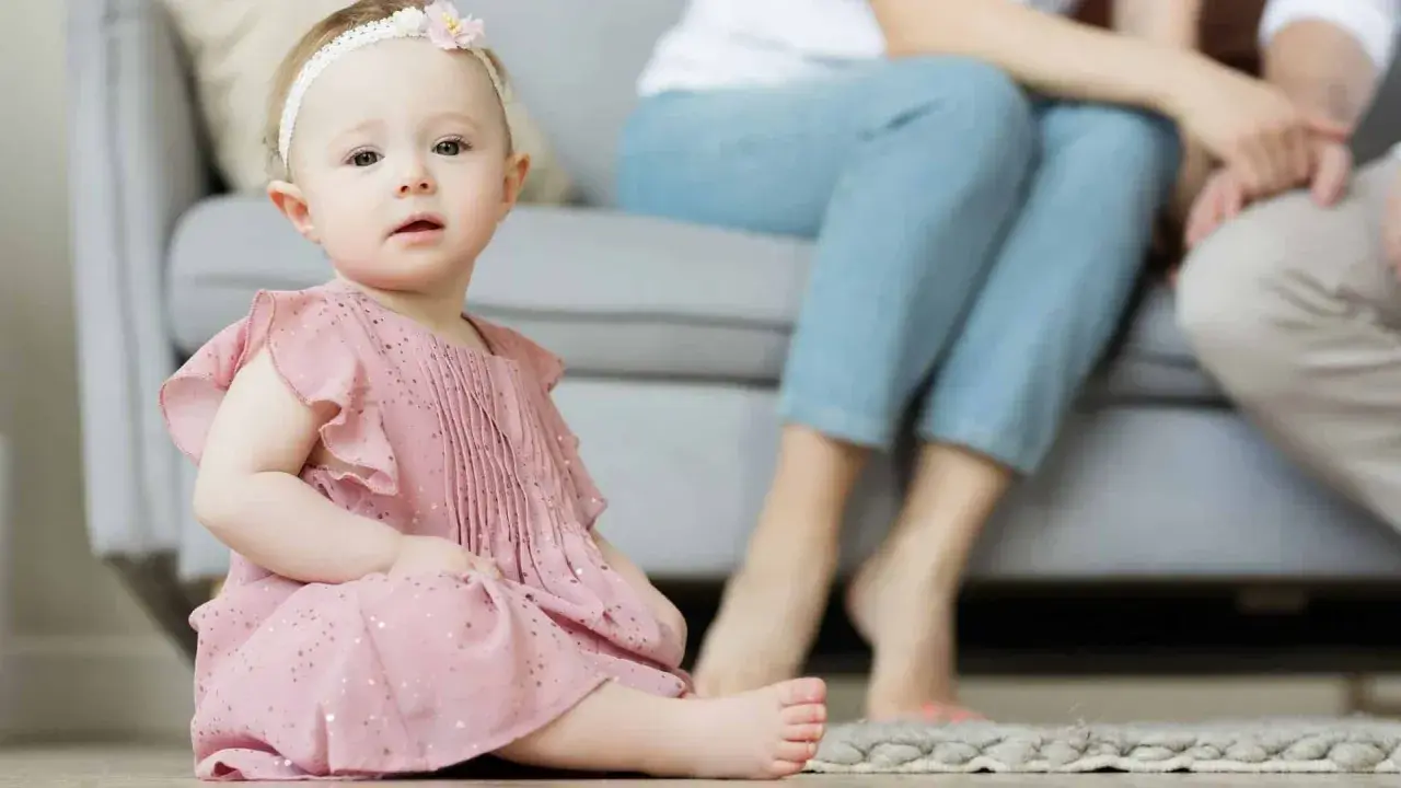 Ein süßes Baby in rosa Kleidchen, mit einem Blumenband im Haar, sitzt auf dem Boden. Im Hintergrund sitzen Eltern. Vielleicht heißt sie Zofia oder Hanna, typisch polnische vornamen weiblich.