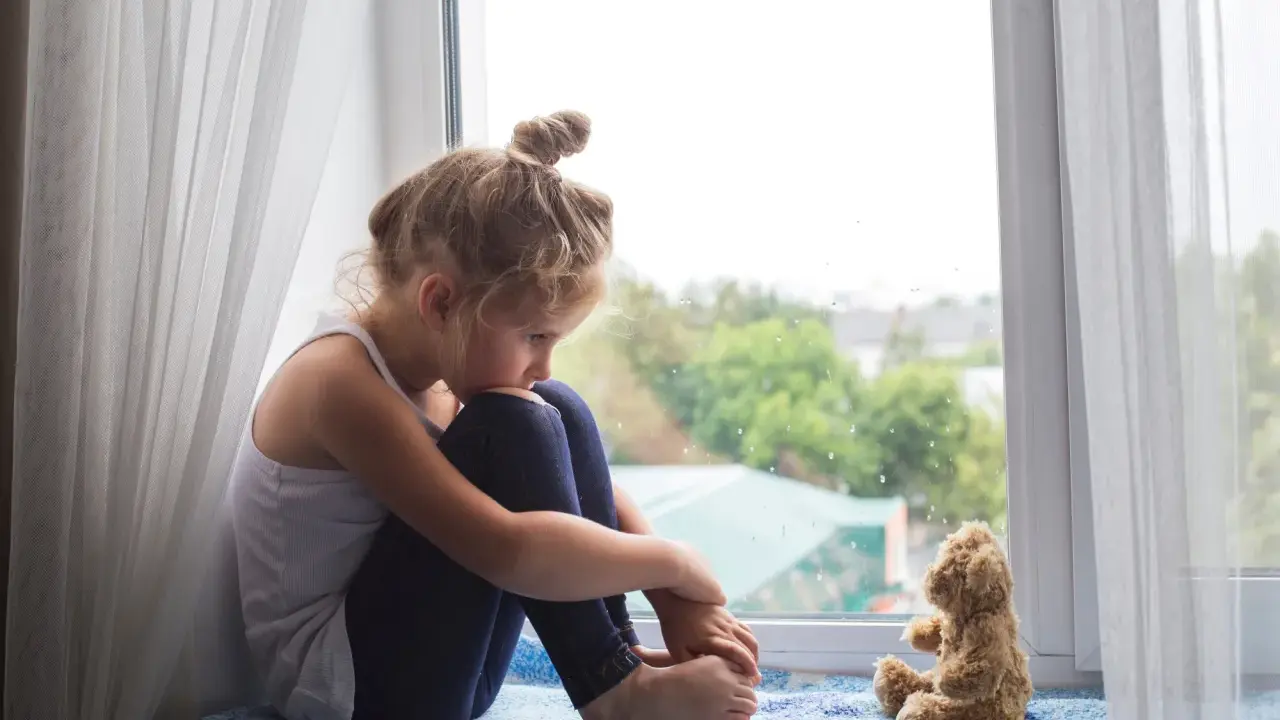 Ein kleines Mädchen sitzt traurig am Fenster, ihr Teddybär neben ihr. Sie denken über den Tod mit Kindern besprechen nach.