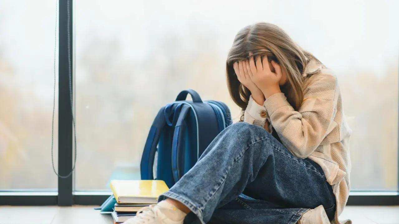 Ein Mädchen mit verdecktem Gesicht sitzt neben einem Rucksack und Büchern. Sie leidet unter **Angst vor Schule ohne Grund**.