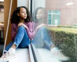 Ein Mädchen sitzt nachdenklich am Fenster. Ihre Reflexion spiegelt den Schmerz des tod eines elternteils im kindesalter wider.