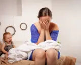 Verzweifelte Mutter mit Baby, das weint. Sie verbirgt ihr Gesicht in den Händen, vielleicht denkt sie: "Regretting motherhood was tun?