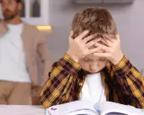 Junge mit Händen am Kopf, der frustriert seine Hausaufgaben macht. Vater im Hintergrund.