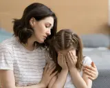 Eine Mutter tröstet ihr weinendes Kind, das sich die Augen zuhält. Möglicherweise kämpft das Kind mit Zwangsgedanken bei Kindern.