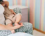 Mutter hält ihr Baby, das auf ihrem Schoß sitzt. Die Streifenwand im Hintergrund ist farbenfroh. Wenn die Mutter während der Stillzeit Durchfall hat, ist das Baby sicher.