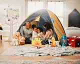 Familie verbringt **gemeinsame Zeit** beim Zelten im Wohnzimmer. Kinder kuscheln mit Teddybären, während Eltern lachen.