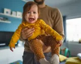 Ein weinendes Baby in gelbem Oberteil und gepunkteter Hose wird von seinem Vater gehalten. Typische "Mama-Phase".