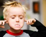 Ein Junge mit blonden Haaren stopft sich Finger in die Ohren, die Augen geschlossen, als ob er nichts hören will. Er verweigert alles.