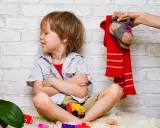Ein Junge sitzt mit verschränkten Armen und einem trotzigen Gesichtsausdruck da. Über ihm hält eine Hand einen roten Schal und ein Paar Socken. Der Junge will sich nicht anziehen.