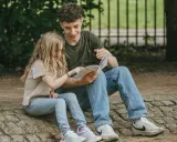Ein junger Mann liest einem Mädchen im Park vor. Trotz der Trennung der Eltern finden die Kinder Trost und Nähe in gemeinsamen Momenten.