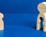 Zwei Holzfiguren auf blauem Grund. Eine kleine Figur steht allein. Die größere Figur hat eine leere Öffnung, die an ein Kind erinnert, und symbolisiert das Bedauern über Mutterschaft.