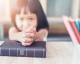 Ein kleines Mädchen betet mit gefalteten Händen über einer Bibel. Vielleicht lernt sie gerade etwas über biblische Frauennamen. Buntstifte liegen daneben.