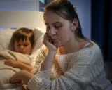 Müde Mutter sitzt neben ihrem schlafenden Sohn, der immer beim Aufwachen weint. Sie hält ihren Kopf in der Hand.
