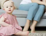 Ein süßes Baby in rosa Kleidchen, mit einem Blumenband im Haar, sitzt auf dem Boden. Im Hintergrund sitzen Eltern. Vielleicht heißt sie Zofia oder Hanna, typisch polnische vornamen weiblich.