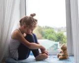 Ein kleines Mädchen sitzt traurig am Fenster, ihr Teddybär neben ihr. Sie denken über den Tod mit Kindern besprechen nach.