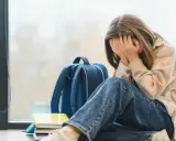 Ein Mädchen mit verdecktem Gesicht sitzt neben einem Rucksack und Büchern. Sie leidet unter **Angst vor Schule ohne Grund**.