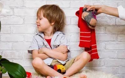 Ein Junge sitzt mit verschränkten Armen und einem trotzigen Gesichtsausdruck da. Über ihm hält eine Hand einen roten Schal und ein Paar Socken. Der Junge will sich nicht anziehen.