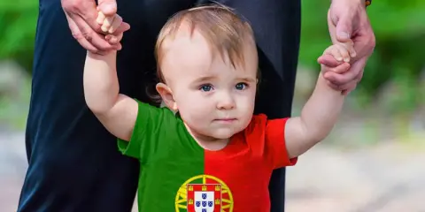 Ein süßes Baby mit portugiesischer Flagge auf dem T-Shirt, gehalten von einer Hand. Vielleicht ein zukünftiger João oder Miguel, inspiriert von portugiesischen Jungennamen.