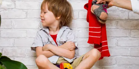 Ein Junge sitzt mit verschränkten Armen und einem trotzigen Gesichtsausdruck da. Über ihm hält eine Hand einen roten Schal und ein Paar Socken. Der Junge will sich nicht anziehen.
