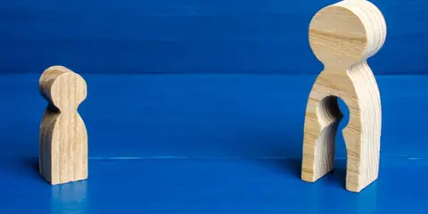 Zwei Holzfiguren auf blauem Grund. Eine kleine Figur steht allein. Die größere Figur hat eine leere Öffnung, die an ein Kind erinnert, und symbolisiert das Bedauern über Mutterschaft.