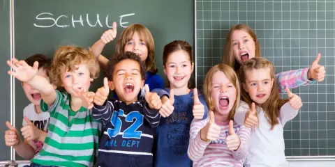 Fröhliche Kinder mit Daumen hoch vor einer Tafel mit dem Wort "SCHULE". Ein perfekter erster schultag!
