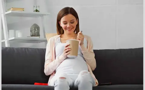 Lächelnde Schwangere isst Eis auf dem Sofa. Ein süßer Genuss während der Eis Schwangerschaft.
