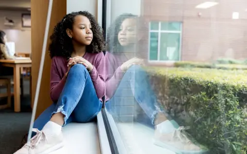 Ein Mädchen sitzt nachdenklich am Fenster. Ihre Reflexion spiegelt den Schmerz des tod eines elternteils im kindesalter wider.