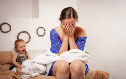 Verzweifelte Mutter mit Baby, das weint. Sie verbirgt ihr Gesicht in den Händen, vielleicht denkt sie: "Regretting motherhood was tun?
