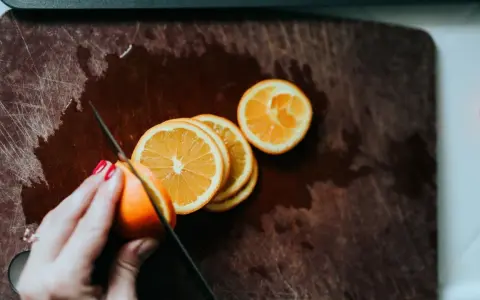 Eine Hand mit roten Nägeln schneidet Orangen auf einem Holzbrett. Orangen in der Schwangerschaft sind eine gesunde Wahl.