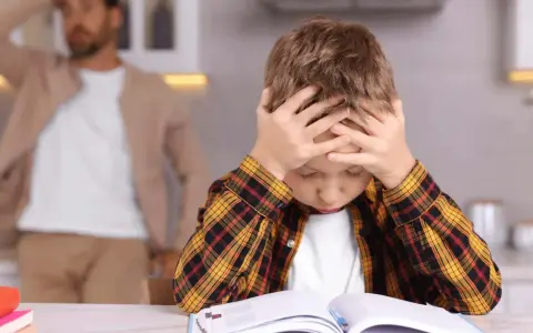 Junge mit Händen am Kopf, der frustriert seine Hausaufgaben macht. Vater im Hintergrund.