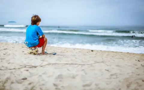 Ein Junge sitzt allein am Strand auf einem Fußball. Er blickt aufs Meer. Meine Tochter 16 hat keine Freunde, sie fühlt sich oft so einsam.