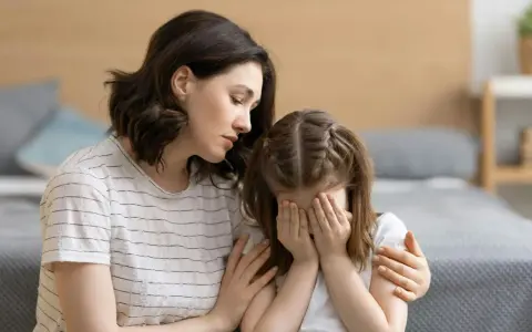 Eine Mutter tröstet ihr weinendes Kind, das sich die Augen zuhält. Möglicherweise kämpft das Kind mit Zwangsgedanken bei Kindern.