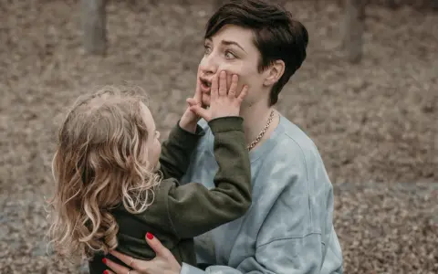 Ein kleines Kind drückt der Mutter spielerisch die Hände ins Gesicht. Vielleicht fragt es sich, warum beleidigen Kinder andere Kinder, wenn sie doch so viel Spaß haben können.