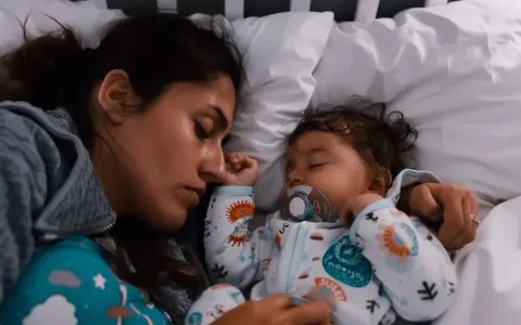 Mutter und Baby schlafen eng aneinander gekuschelt im Bett. Das Baby hat einen Schnuller im Mund. Ein friedlicher Moment, der zeigt, dass man nicht immer alleine ins Bett gehen muss.