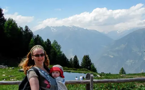 Frau mit Baby im Tragetuch genießt die Berglandschaft. Ein Beispiel für alleine reisen mit Kind.