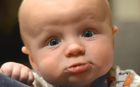 Nahaufnahme eines Babys mit großen blauen Augen und einem lustigen Gesichtsausdruck. Das Baby scheint sich selbst zu erkunden, seine Hand ruht auf einer dunklen Oberfläche.