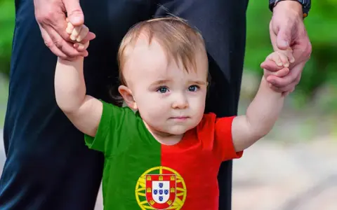 Ein süßes Baby mit portugiesischer Flagge auf dem T-Shirt, gehalten von einer Hand. Vielleicht ein zukünftiger João oder Miguel, inspiriert von portugiesischen Jungennamen.