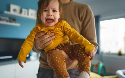 Ein weinendes Baby in gelbem Oberteil und gepunkteter Hose wird von seinem Vater gehalten. Typische "Mama-Phase".