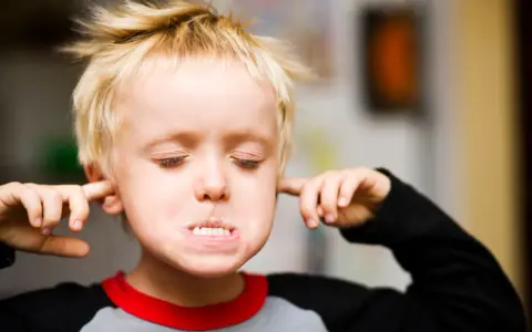 Ein Junge mit blonden Haaren stopft sich Finger in die Ohren, die Augen geschlossen, als ob er nichts hören will. Er verweigert alles.
